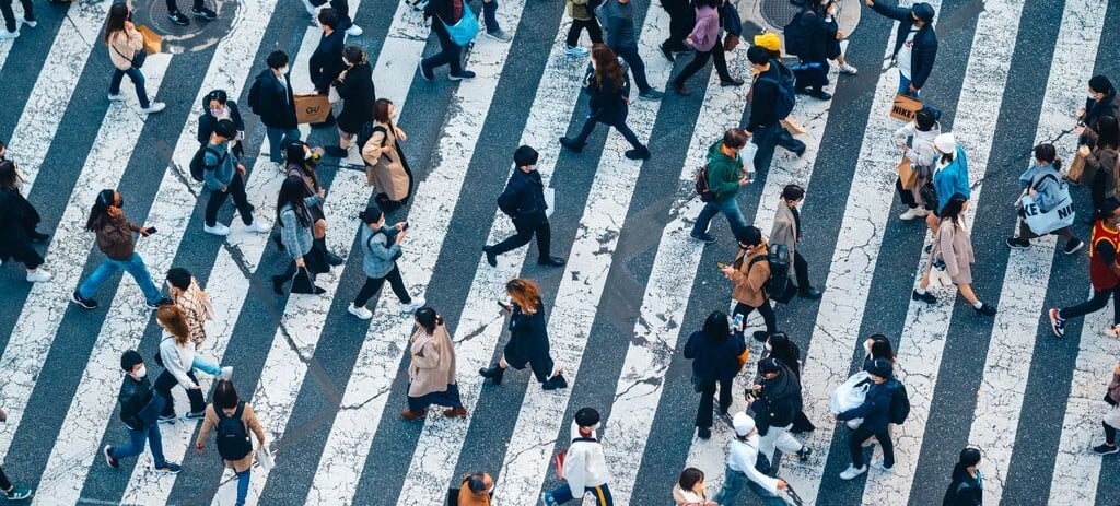 Overhead view of a busy pedestrian crosswalk with people walking in multiple directions on wide white stripes in an urban city setting.