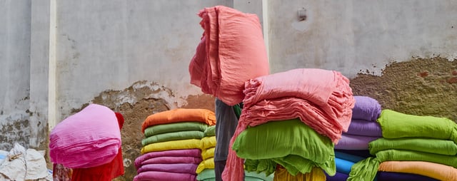 Pilas de fardos de tela de colores vivos —rosado, verde, naranjo, morado y azul— frente a un muro de hormigón desgastado